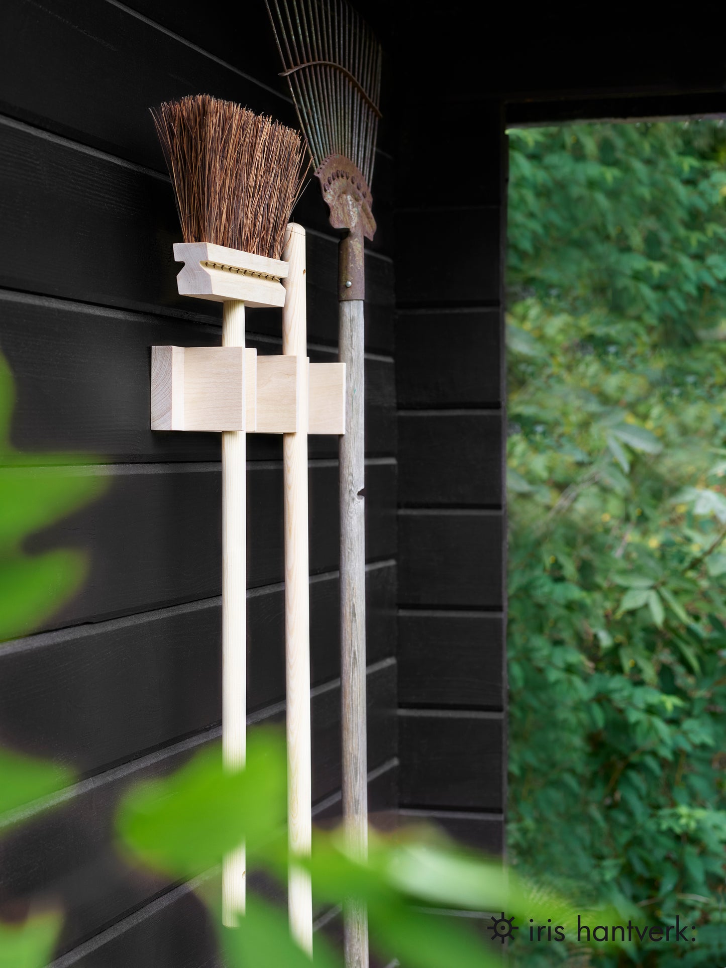 Wooden broom hanging off a black building with other tools next to it.