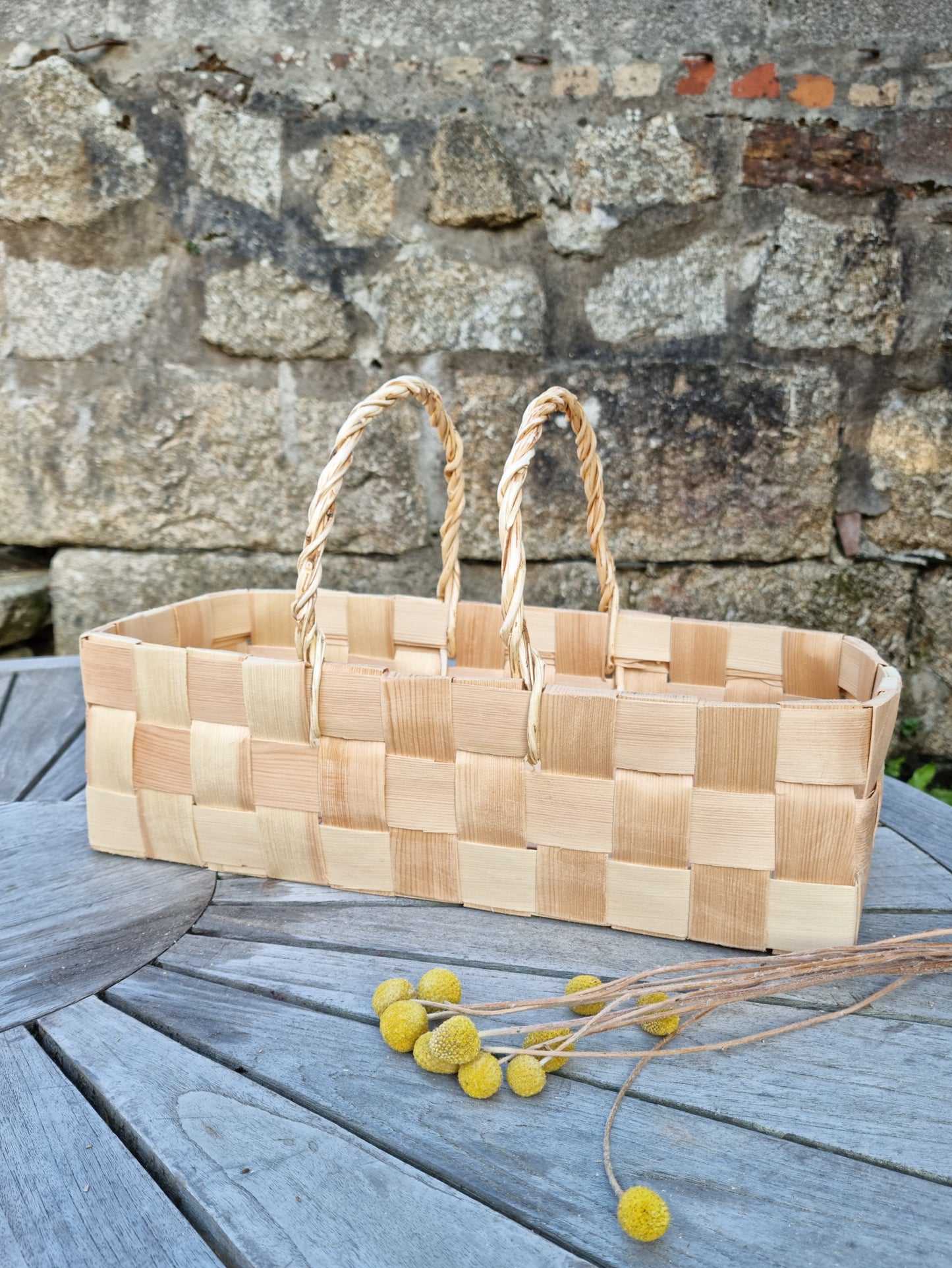 Woven basket placed in front of a brick wall on a wooden garden table. Yellow flowers are placed next to it on the table.