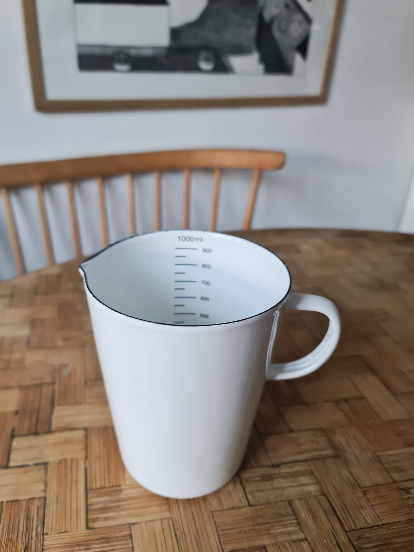 A white enamel measuring jug with black markings and a handle, displayed on a wooden table with a wooden chair in the background.