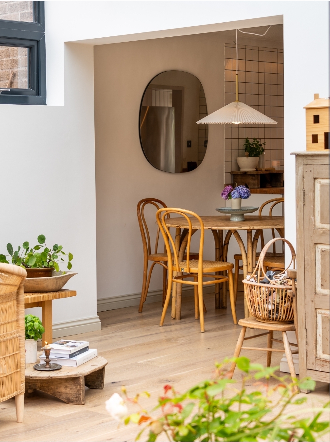 Warm, inviting dining nook with bentwood chairs and rustic wooden table, styled with fresh hydrangeas. A pleated pendant light and organic mirror add soft design details, with natural textures and plants completing the calm, Scandinavian-inspired space.
