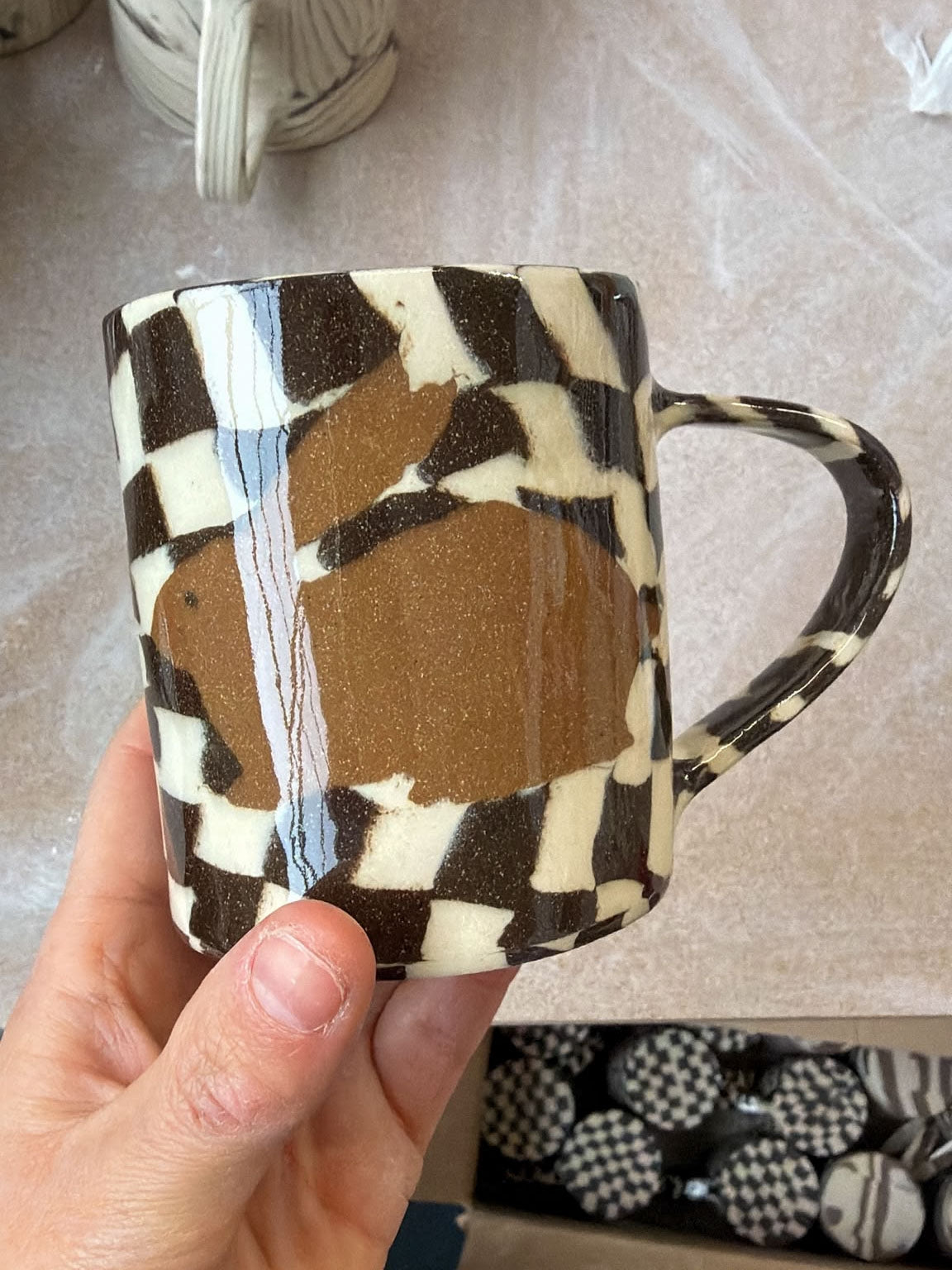 A hand holding a ceramic mug with a brown and cream checkerboard pattern and a large brown rabbit silhouette on the front, set against a neutral tabletop background.