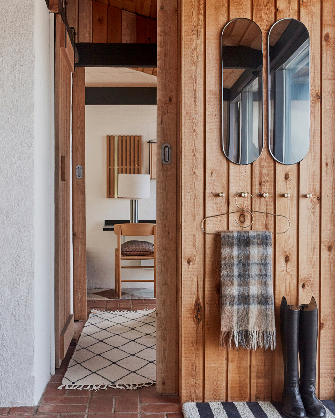 A rustic wooden entryway with two oval mirrors, a plaid throw towel on a hanger, black boots, and a doorway leading into a home interior.