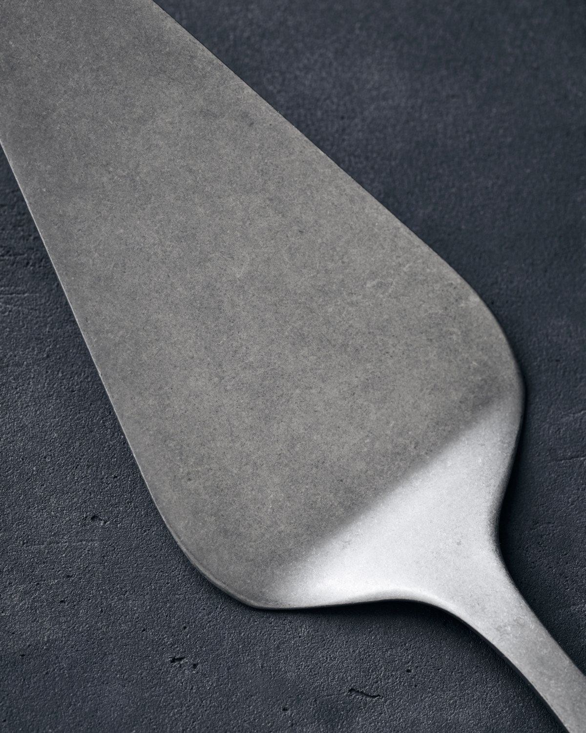 A brushed stainless steel cake server with a smooth, silver finish placed on a dark textured surface.
