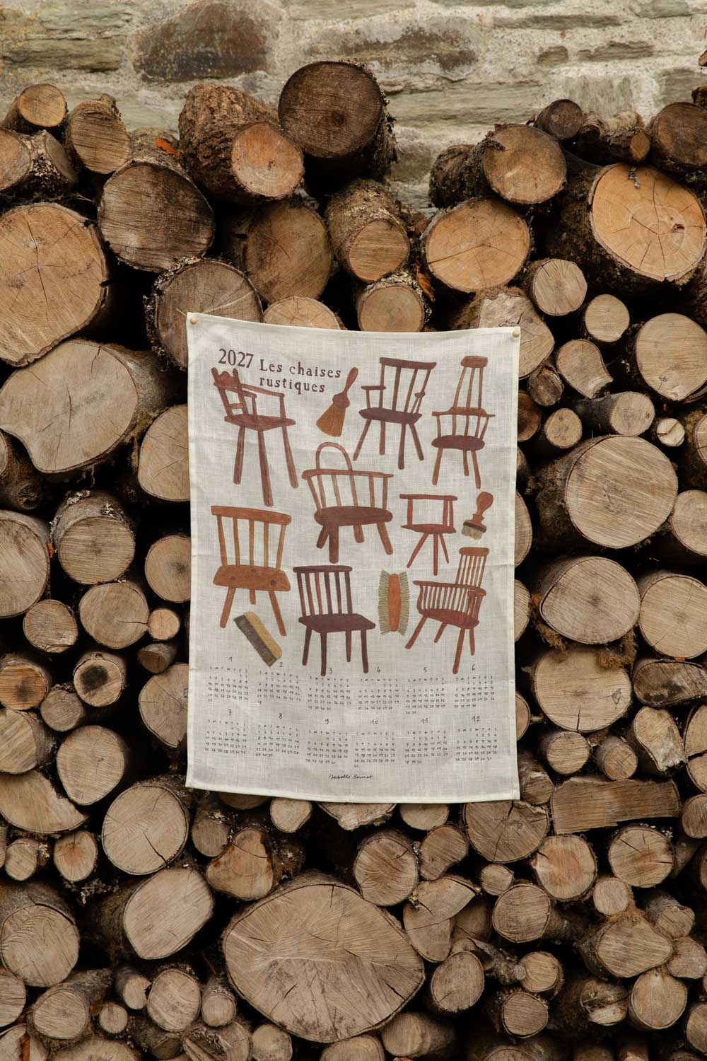 2027 Les Chaises Rustiques linen tea towel calendar pinned to a stack of logs