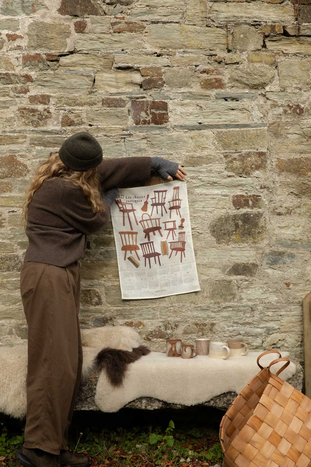 Woman pinning a rustic chairs linen tea towel calendar to a stone wall above a sheepskin bench