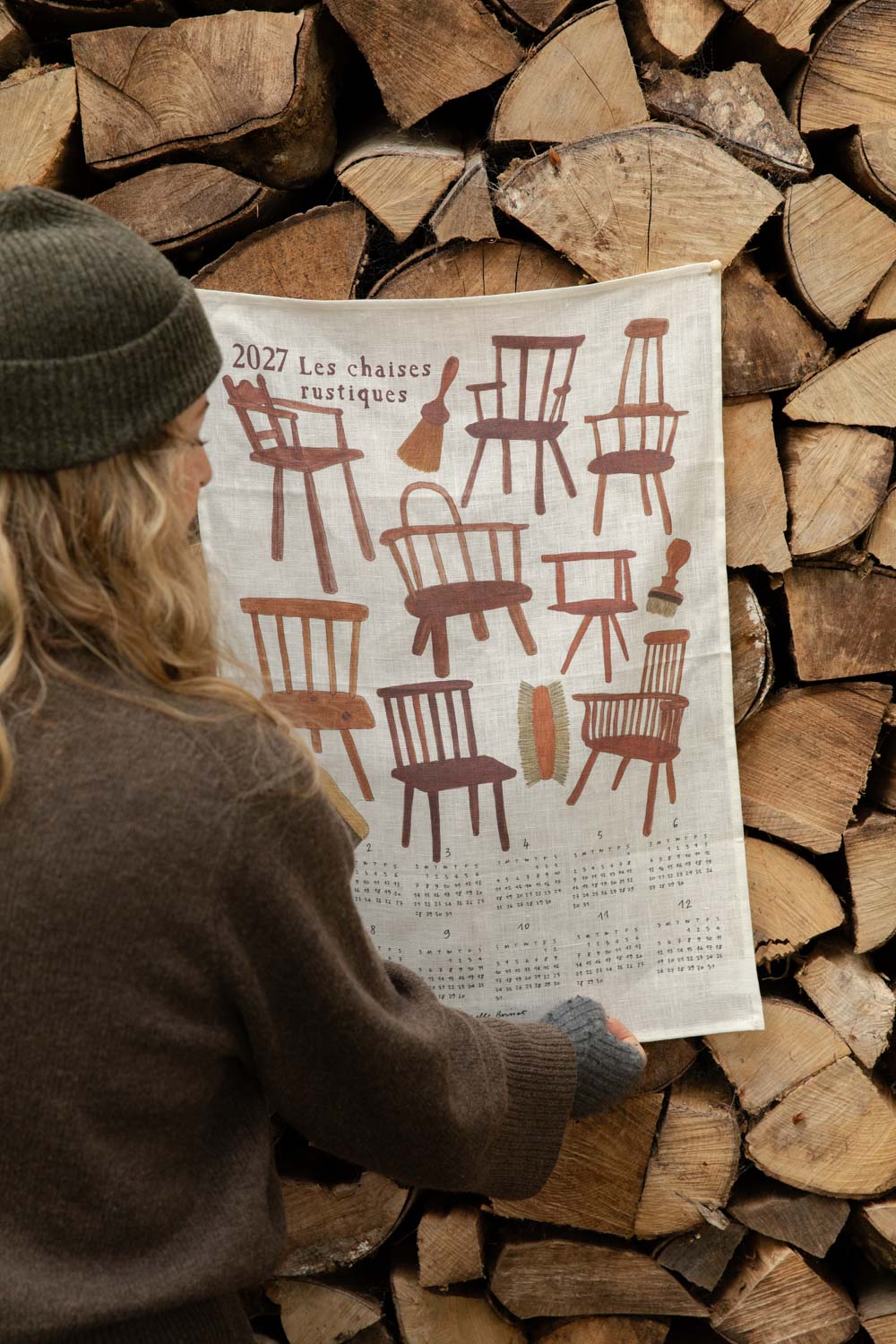 Woman in a Granite Heather wool beanie holding a 2027 linen tea towel calendar printed with rustic chairs against a log pile