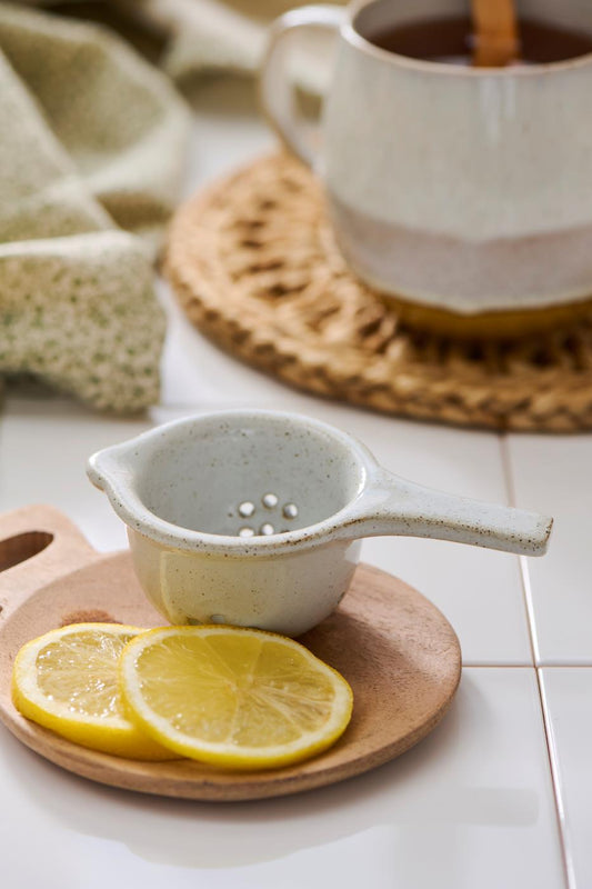 Porcelain tea strainer with speckled glaze, resting on a wooden board with two lemon slices, a ceramic mug and woven coaster blurred in the background. Perfect for straining tea or lemon juice in a calm, minimal kitchen setting.