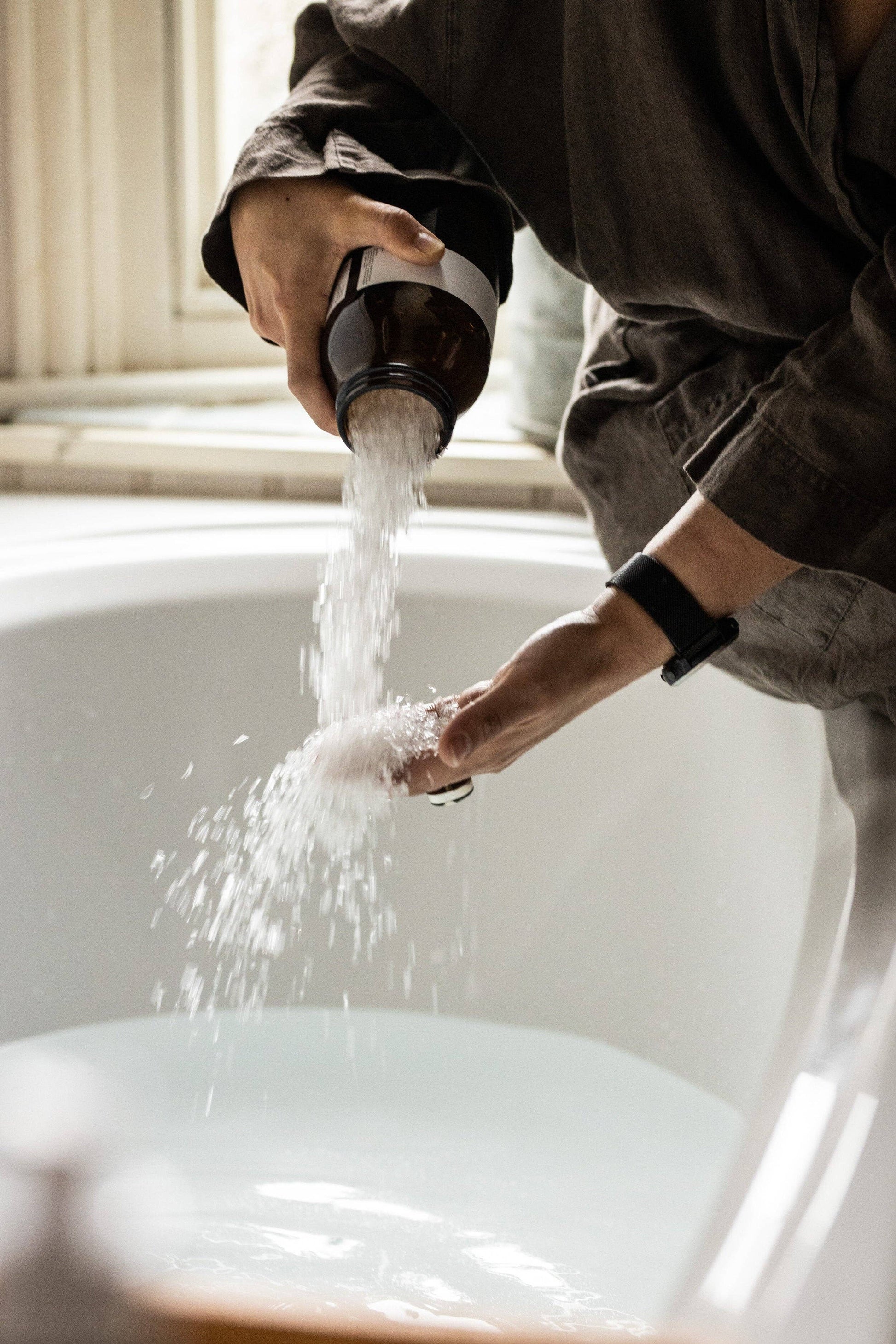 Hands pouring Harvest bathing salts from amber jar into white bath