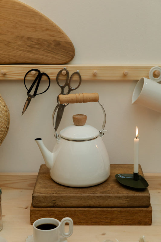 White kettle with wooden handle on a stack of wooden boards, surrounded by kitchen items.