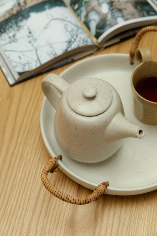 Ceramic teapot on ceramic tray with wicker handles, next to a cup of tea on a wooden surface.