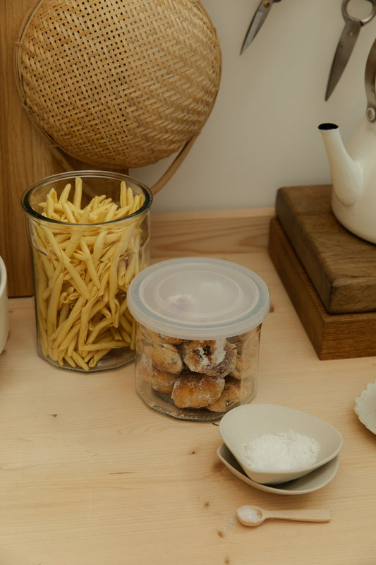 Glass jars with snacks on a wooden table, with a teapot and woven basket in the background.