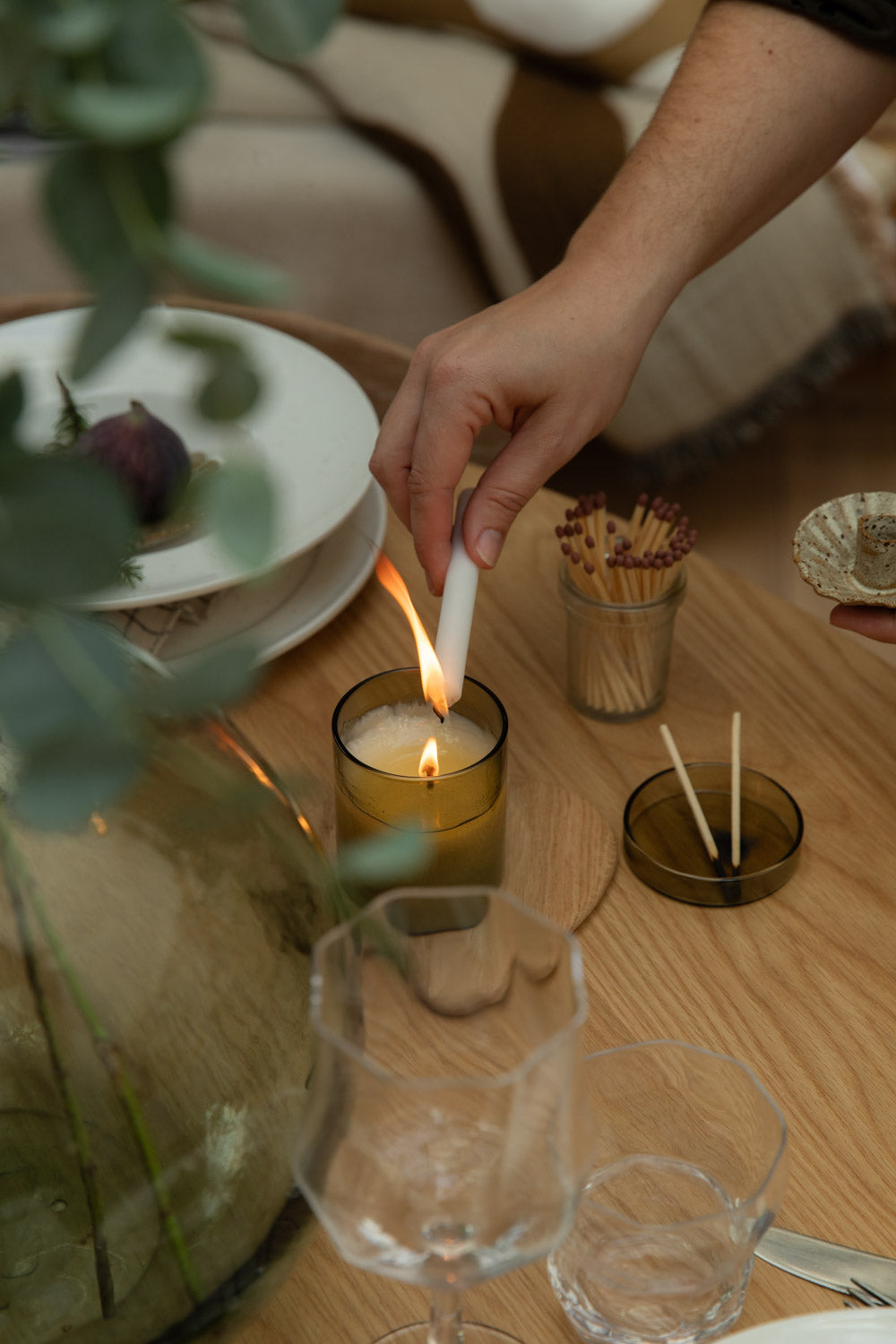 Person lighting a candle on a wooden table with glasses, plates, and a vase.