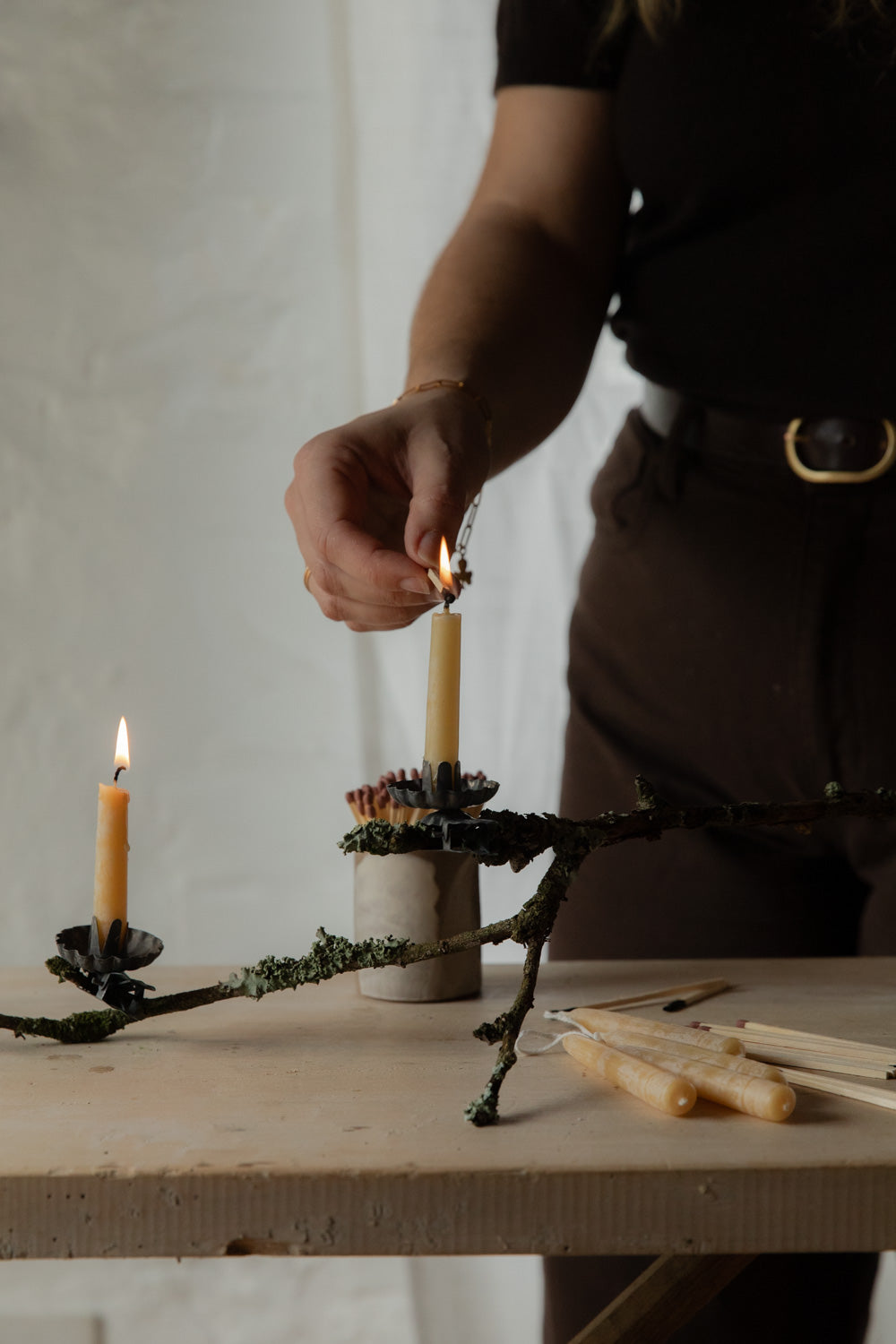 Person lighting a candle on a decorative stand with plants and tools on a wooden surface.