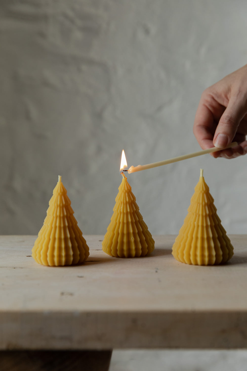 Three cone-shaped beeswax candles on a wooden surface with a hand lighting one of them.