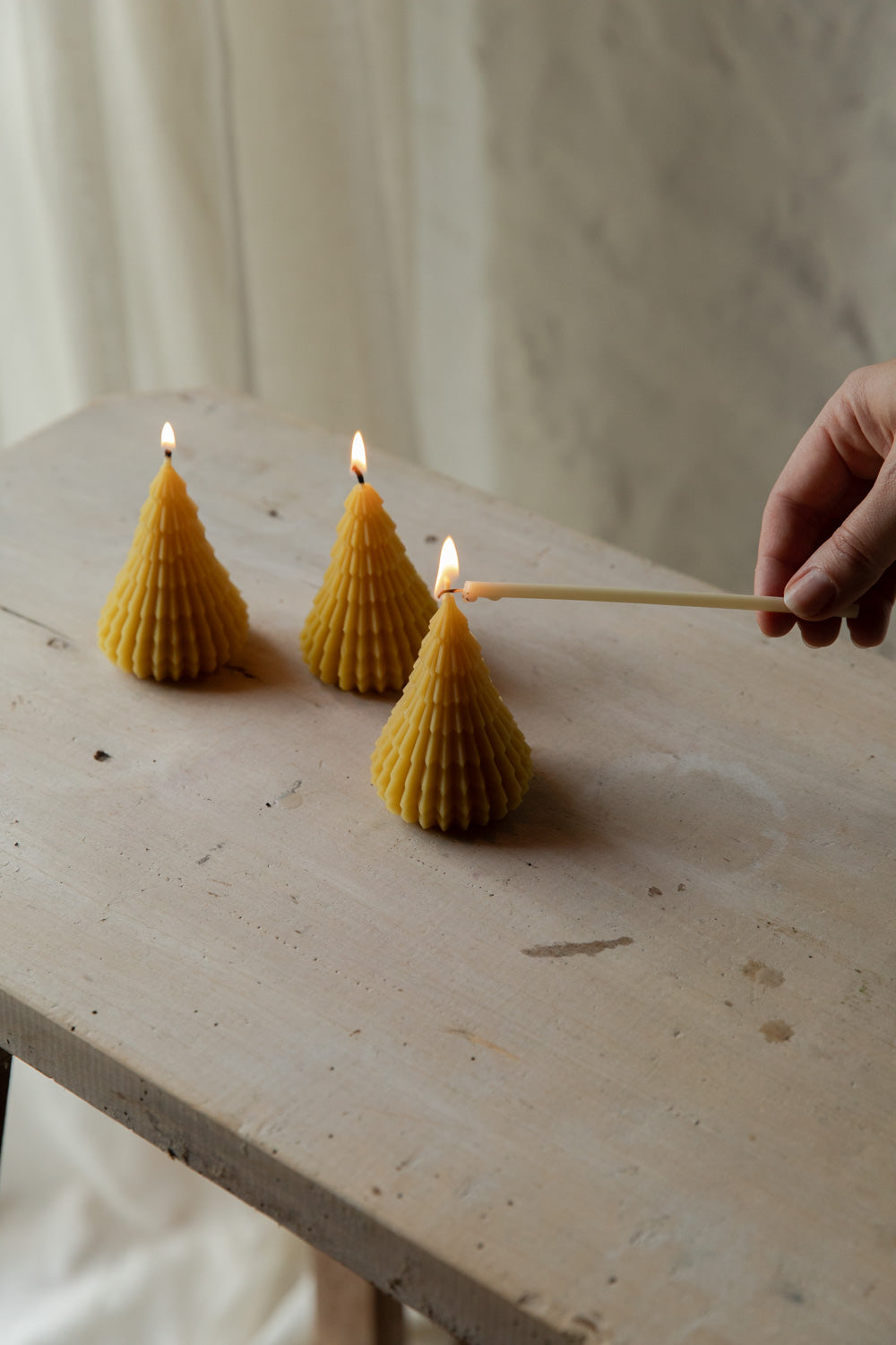 Three beeswax tree-shaped candles being lit with a match on a wooden surface.