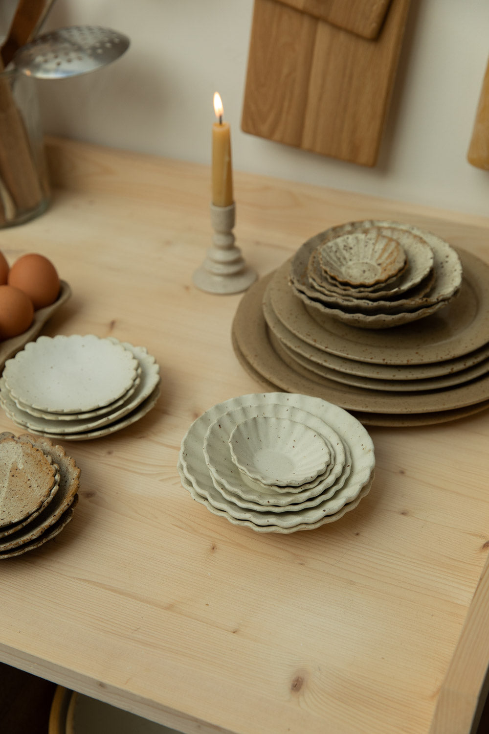 Stacks of ceramic plates on a wooden table with a candle and eggs in the background.