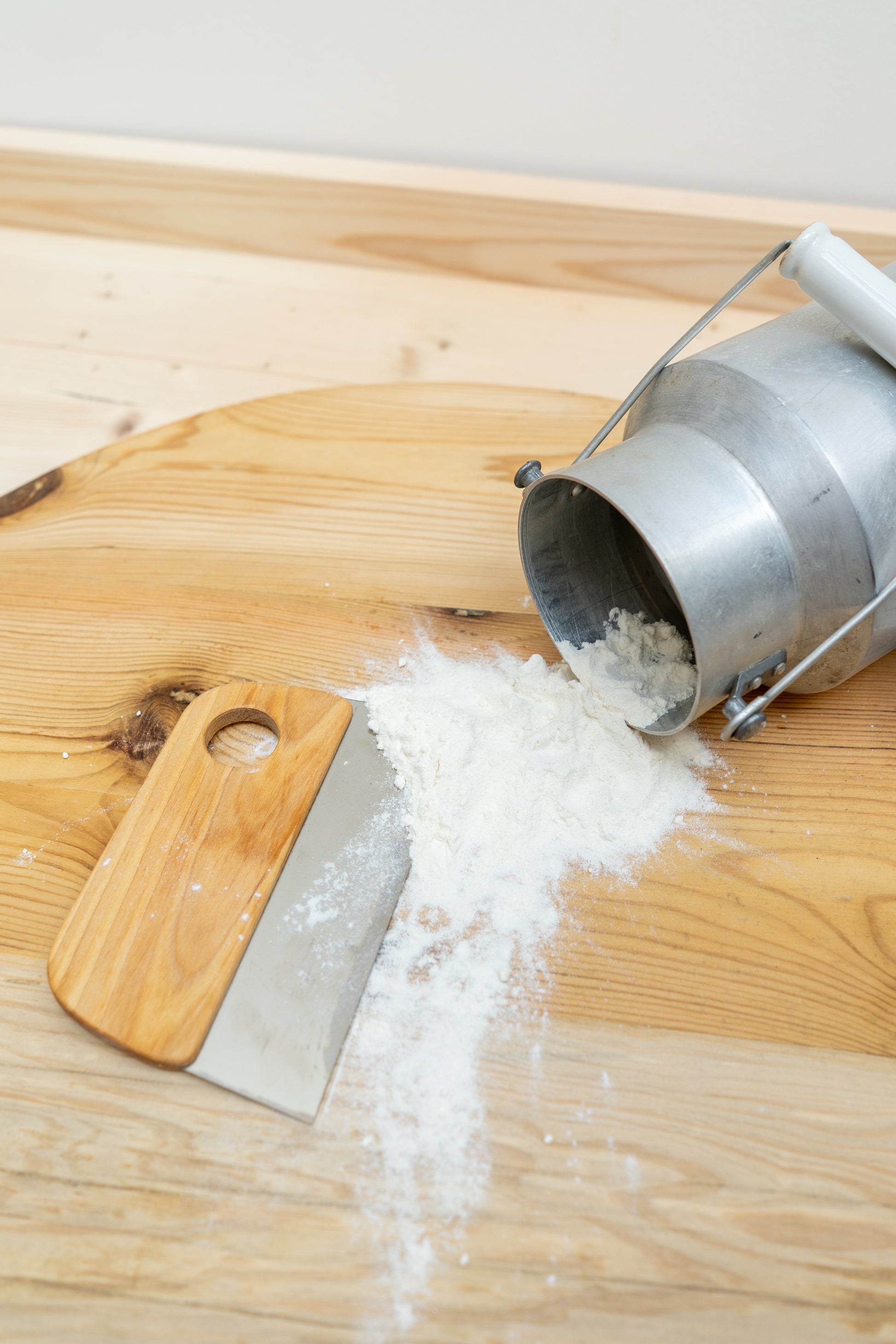 Wooden cutting board with a metal and wood dough scrapper, a pile of flour and metal milk pale on a light background
