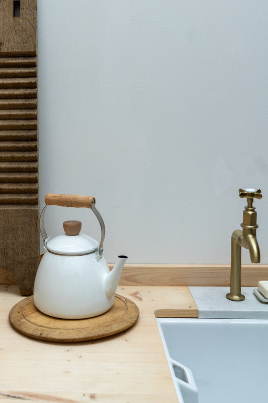 White teapot with wooden handle on a wooden coaster next to a sink with brass faucet.