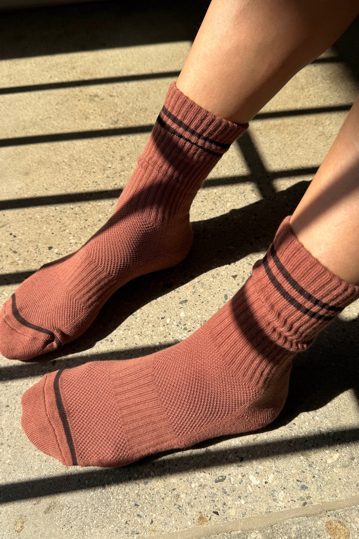 A close-up of a person's feet wearing ribbed crew socks in a dusty clay color with thin black stripes near the cuffs and toes, standing on a concrete surface in direct sunlight.
