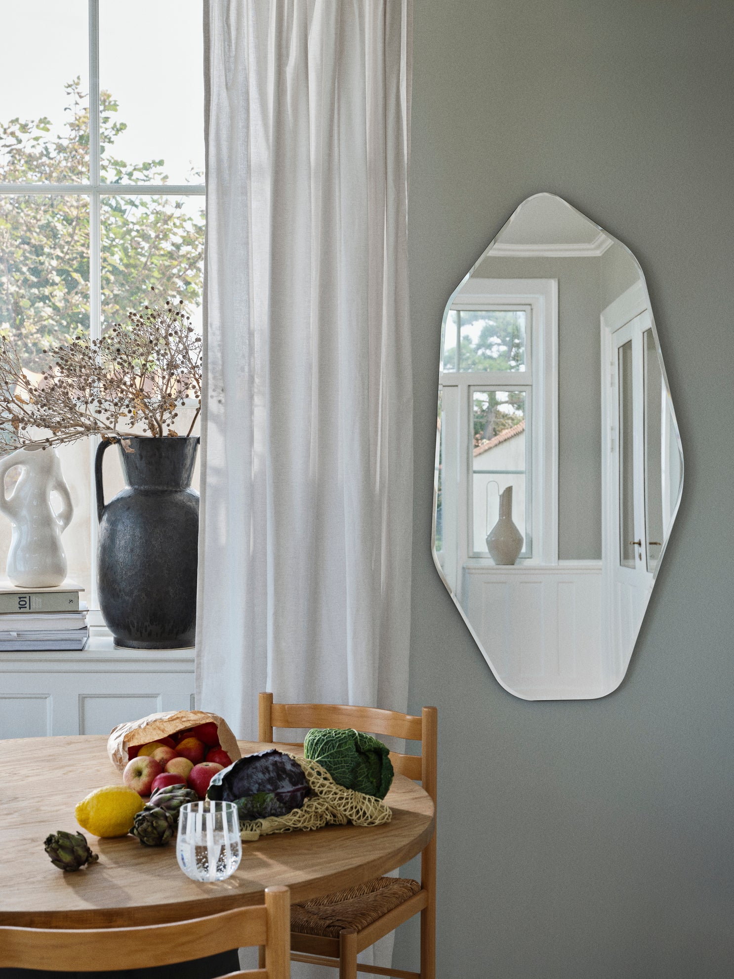 Dining area with a mirror on the wall, table with fruits and a vase, and window with white curtains.