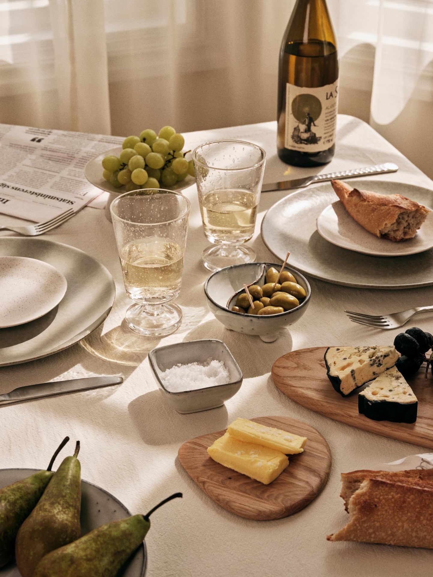 Beautifully laid table, natural cloth, cheese, olives, glassware and ceramics.