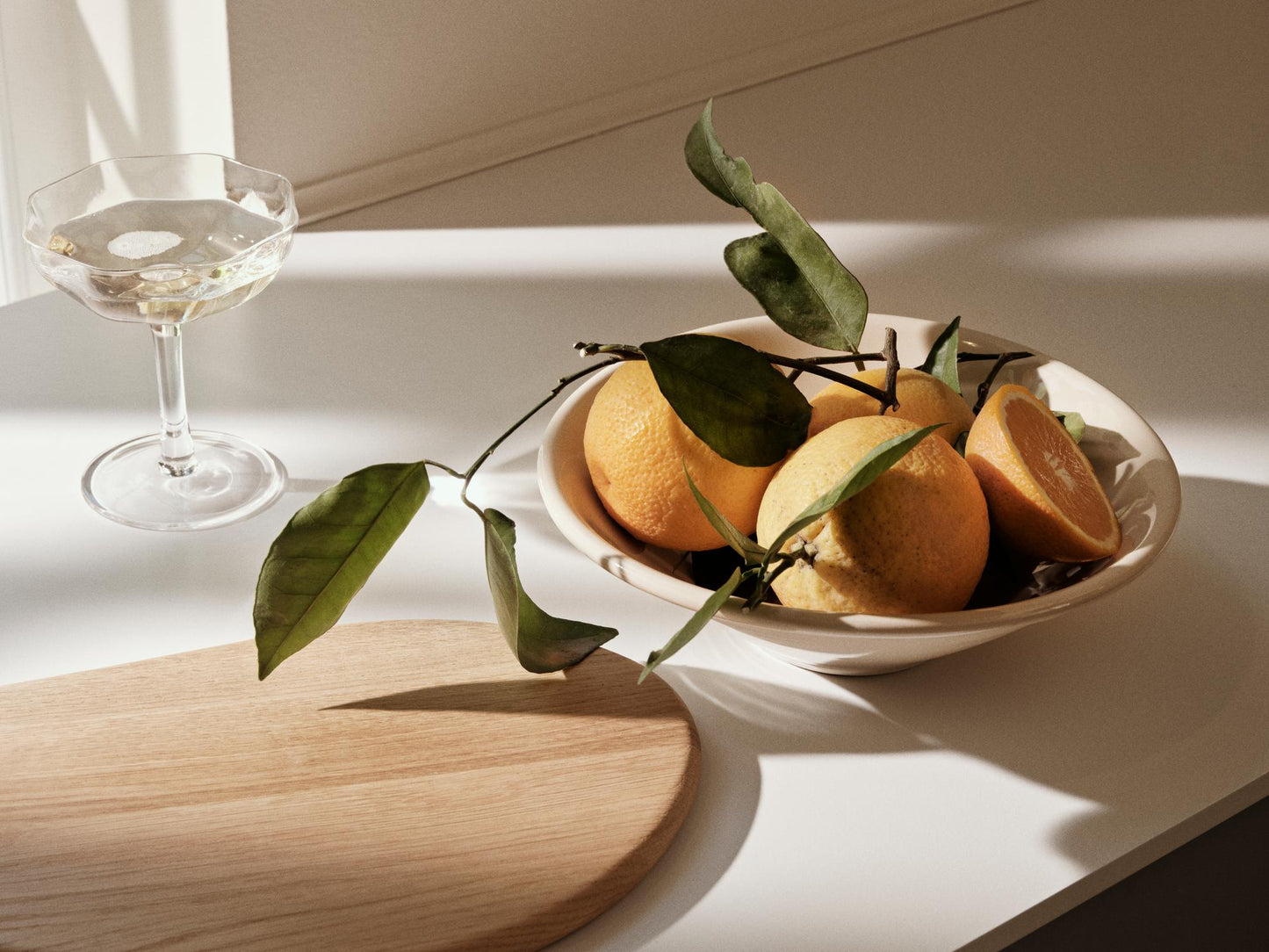 Bowl of oranges with leaves on a table next to a glass of white wine.