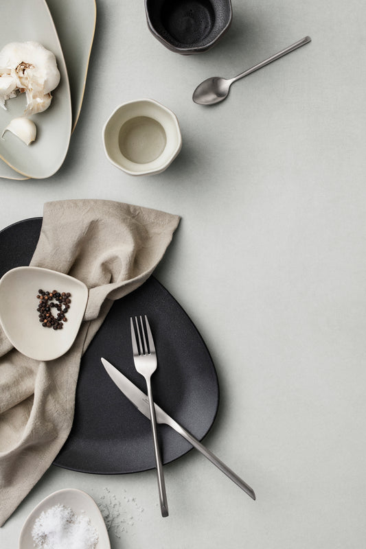 Table setting with black plates, silver cutlery, and neutral napkin on a light gray surface.