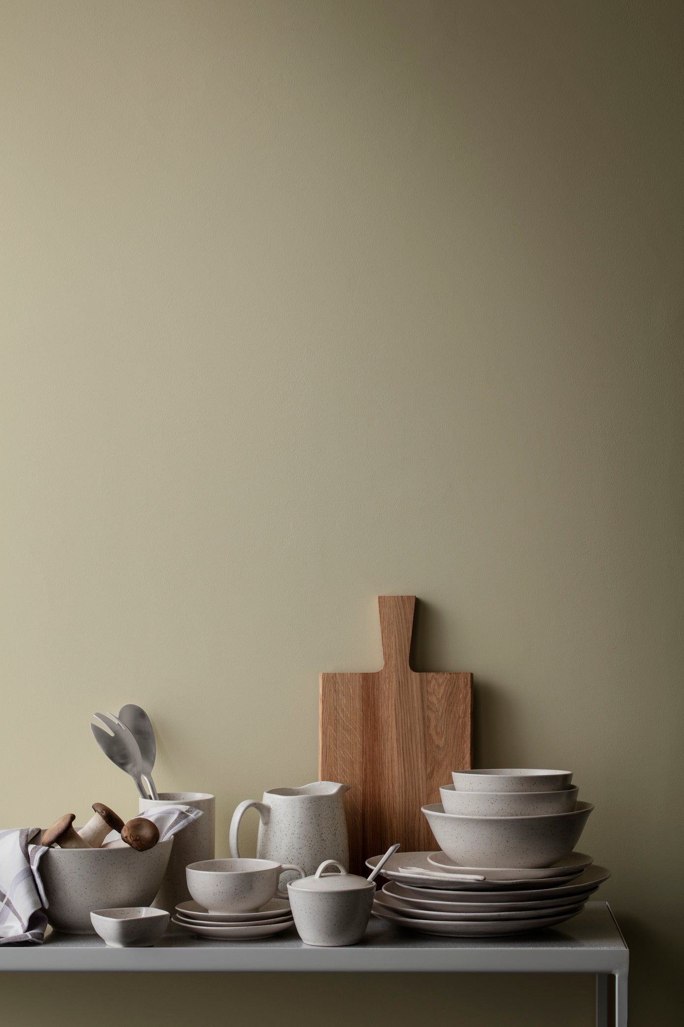Set of ceramic dishes and a wooden cutting board on a table against a green wall.