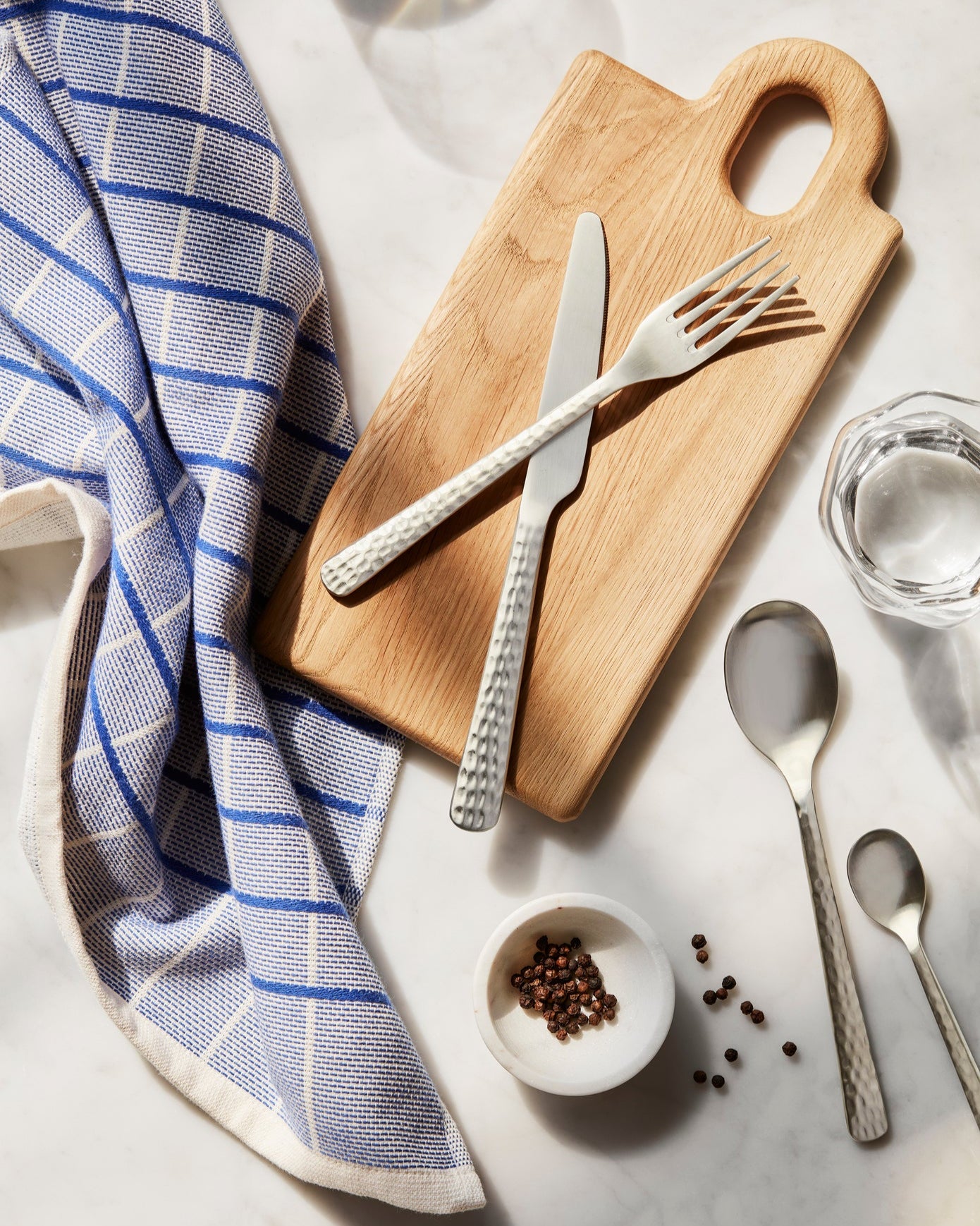 Wooden cutting board with silverware, a towel, and small bowl of peppercorns on a marble surface.