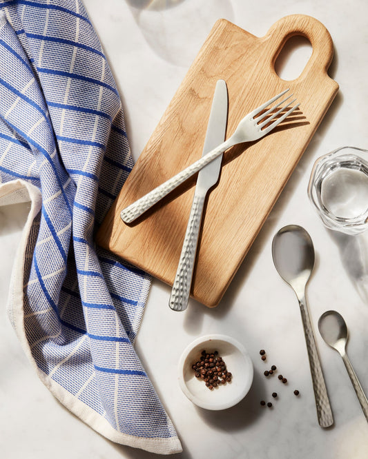 Wooden cutting board with silverware, a towel, and small bowl of peppercorns on a marble surface.