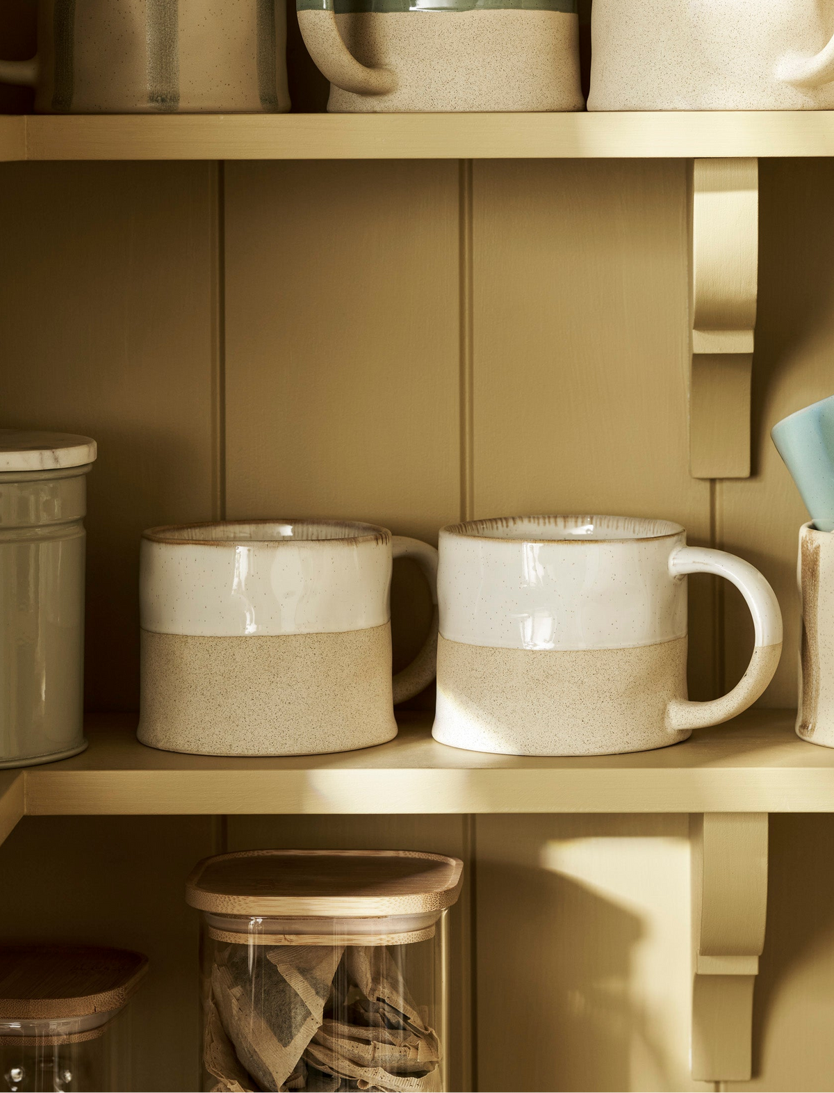 Two ceramic mugs with a two-tone design of glossy cream on the top half and speckled beige on the bottom half, displayed on a wooden shelf alongside other homeware.