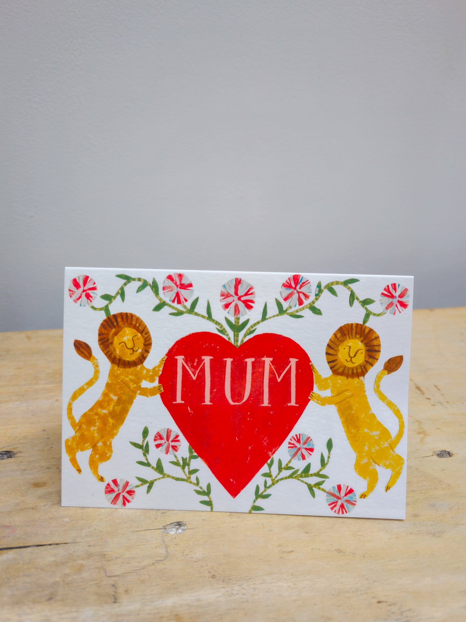Card with two lions holding a red heart with 'MUM' text on a wooden surface.
