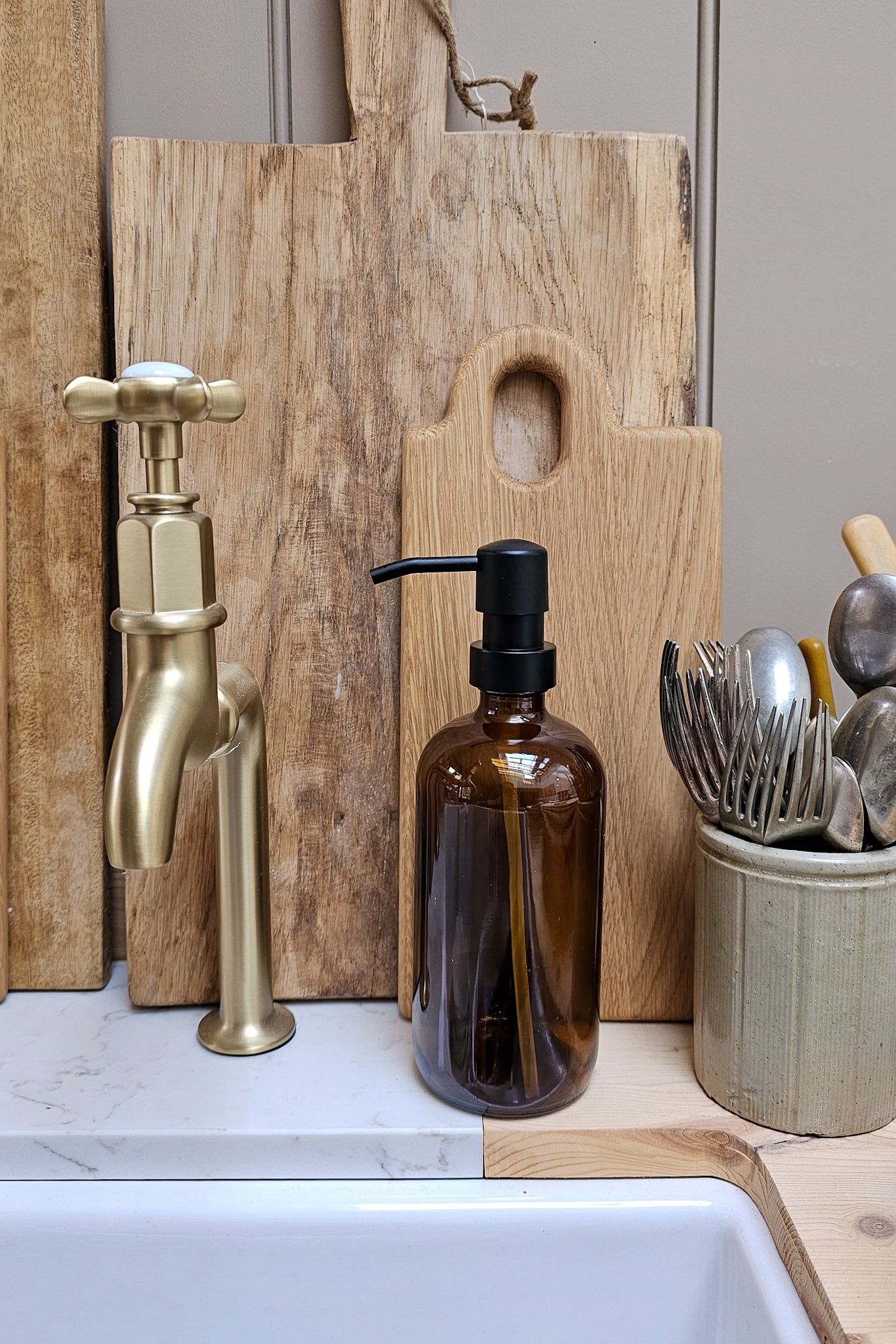 Amber glass soap dispenser bottle with matte black pump on a marble worktop, styled beside a brass kitchen tap, wooden chopping boards and a ribbed ceramic utensil holder.