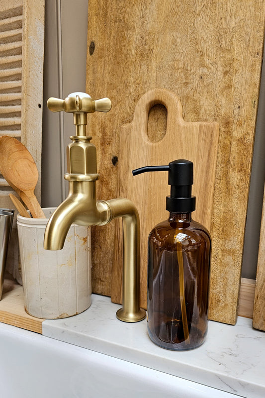 Amber glass refillable soap dispenser bottle with matte black pump, styled on a marble kitchen worktop beside a brushed brass tap and wooden utensil pot.