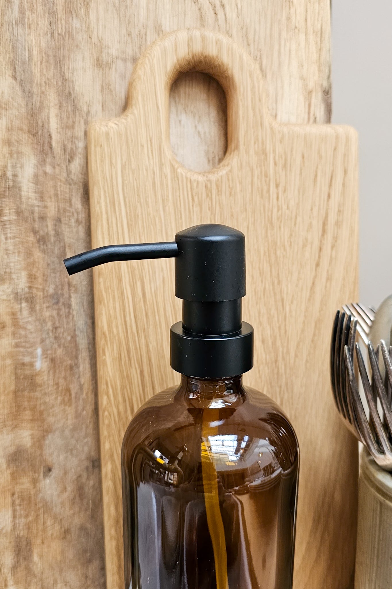 Close-up of the matte black pump on an amber glass soap dispenser bottle, with a wooden chopping board and cutlery pot in the background.