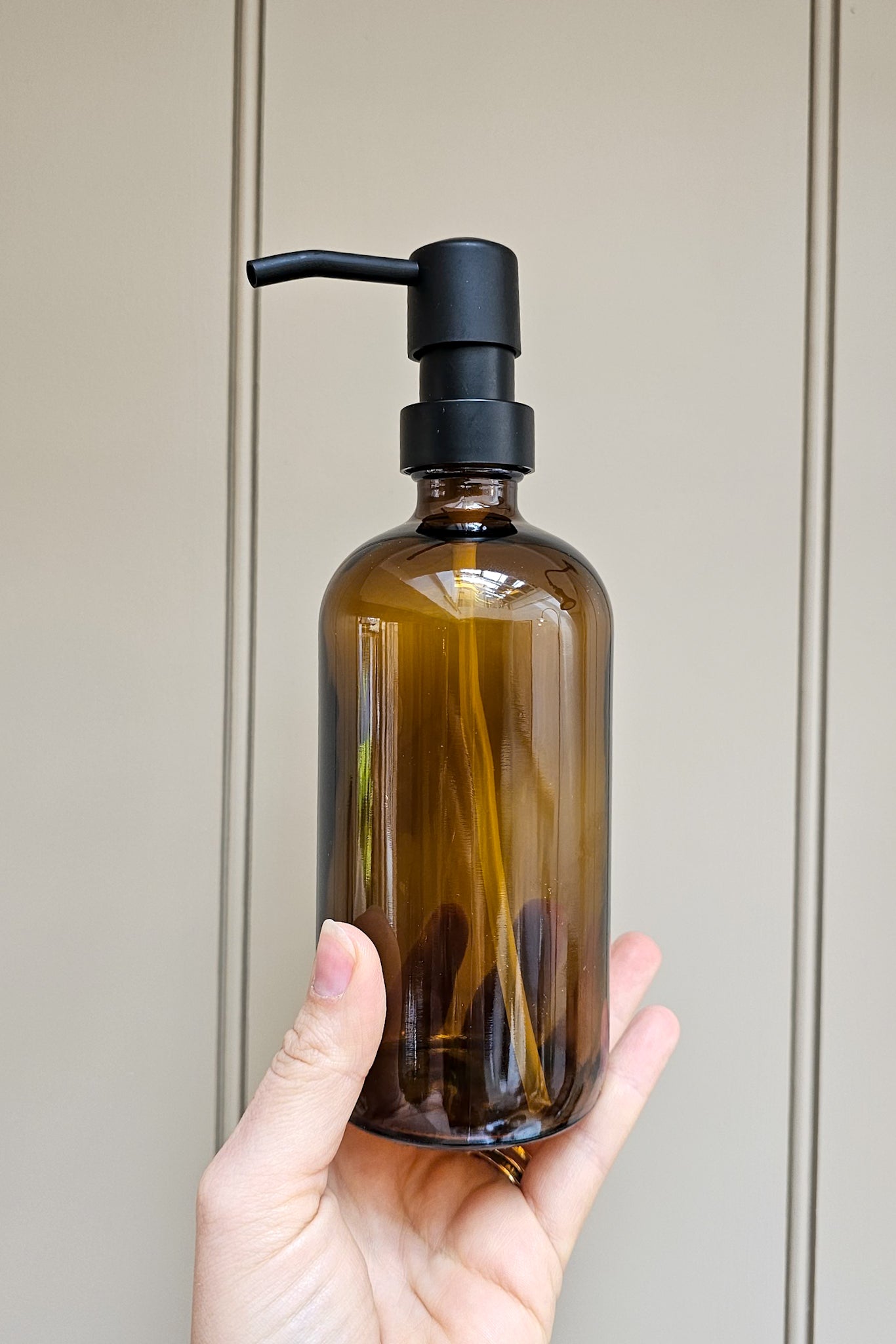 Hand holding an amber glass refillable soap dispenser bottle with matte black pump against a panelled grey-green wall.