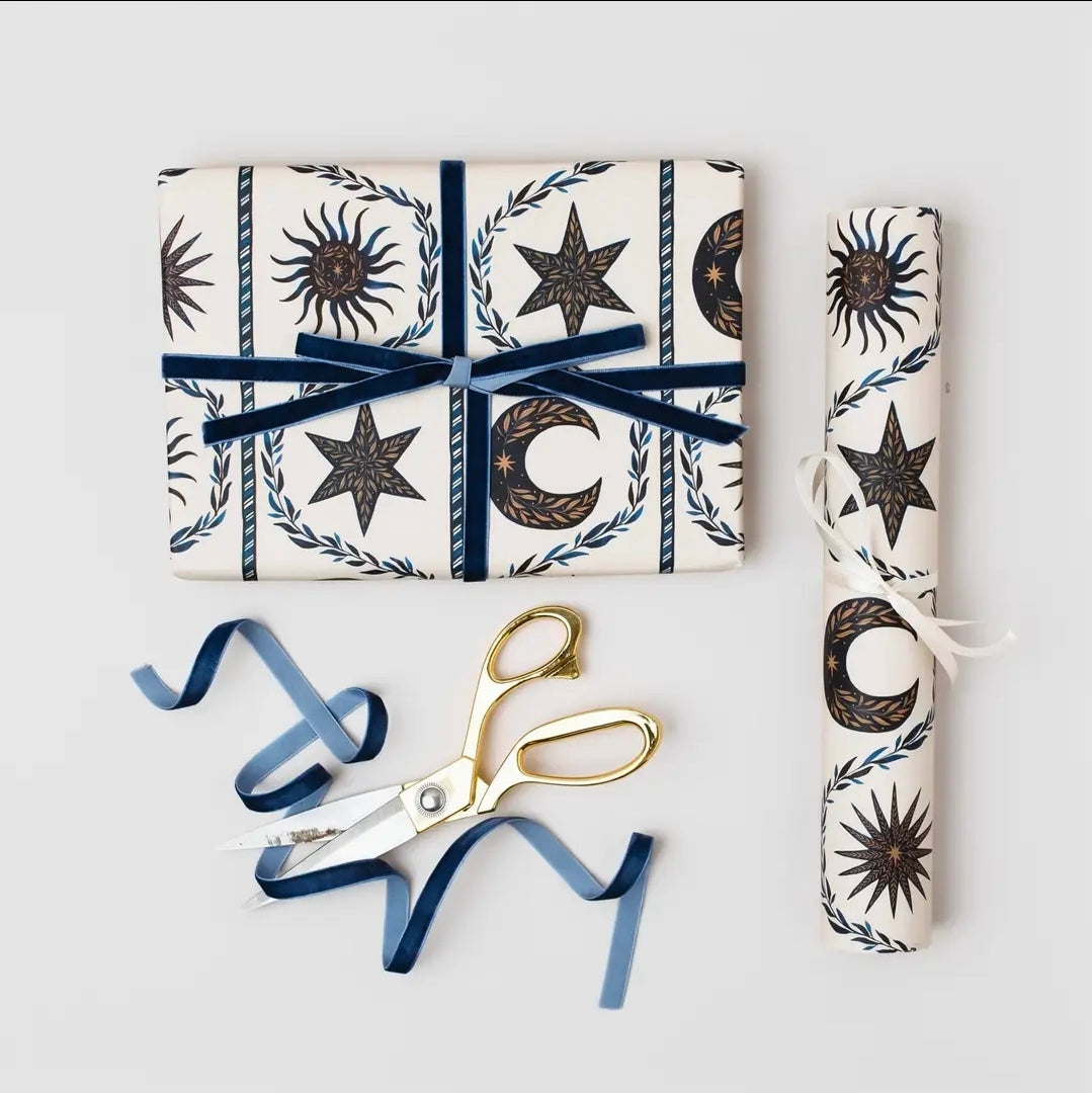 Present wrapped in celestial gift wrap and tied with navy velvet ribbon, beside a rolled sheet, gold scissors and blue ribbon