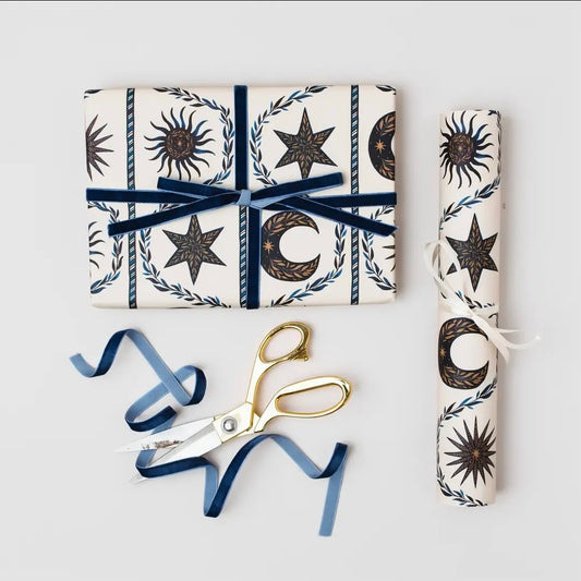 Present wrapped in celestial gift wrap and tied with navy velvet ribbon, beside a rolled sheet, gold scissors and blue ribbon