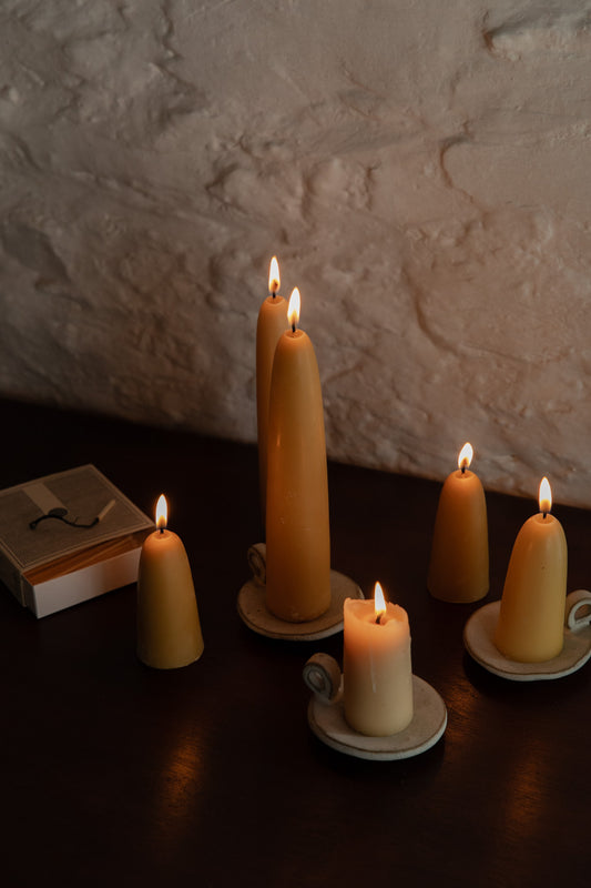 Lit beeswax candles of different heights on handmade ceramic candle plates against a whitewashed brick wall