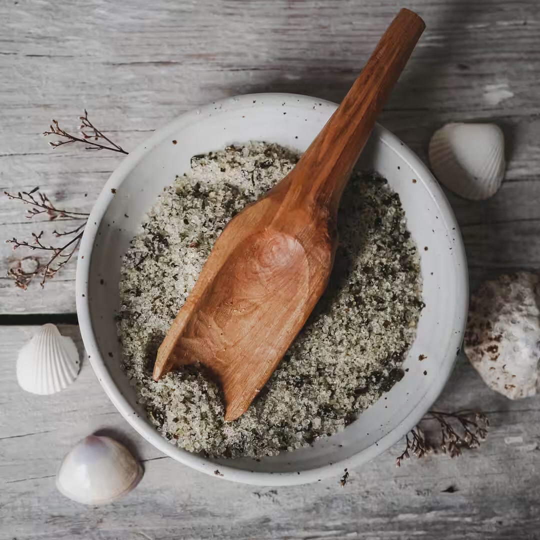 Seaweed bath salts flecked with dried seaweed in a speckled ceramic bowl with a wooden scoop