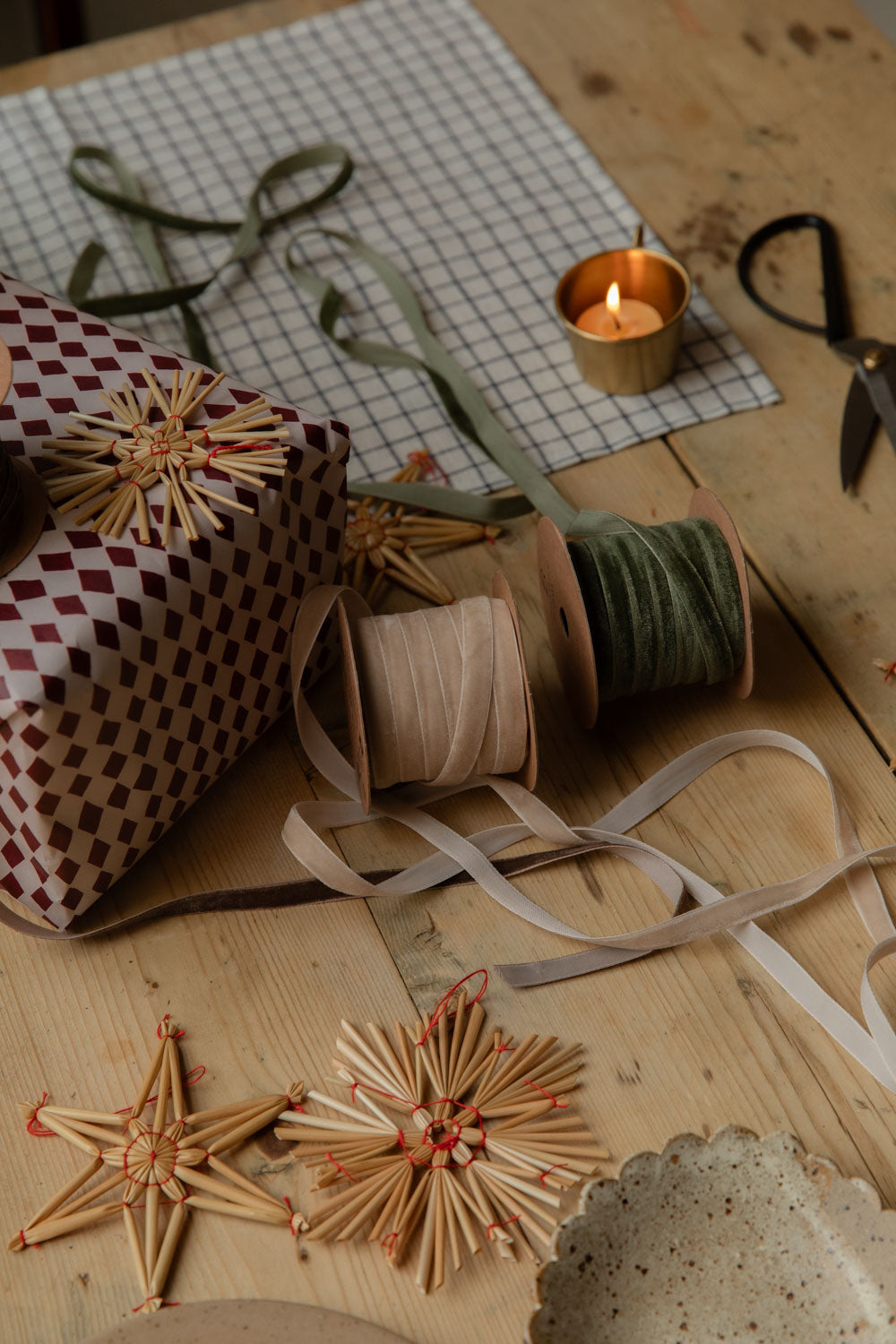 Velvet ribbon spools, gingham wrapping paper, straw stars and a lit candle on a wooden table