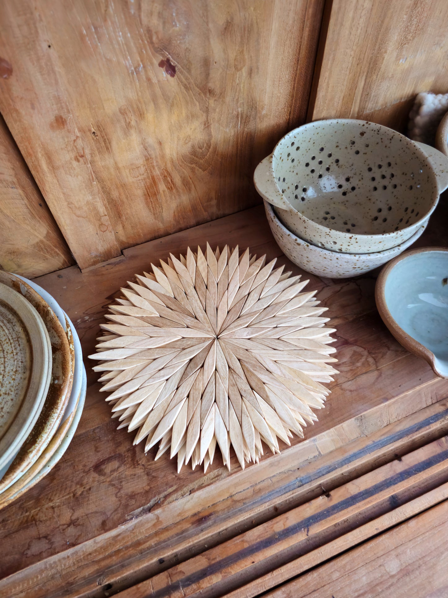 Large natural wooden starburst trivet on a wooden kitchen shelf, styled with handmade ceramic bowls and plates