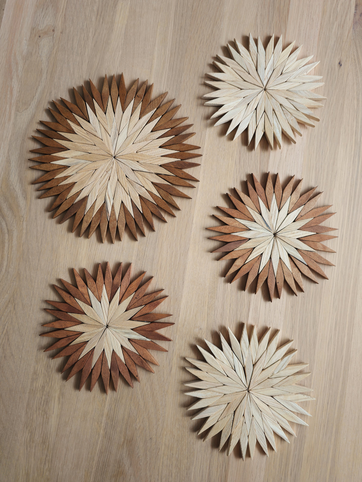 Five handmade wooden starburst trivets in natural and two-tone colourways, small and large sizes, on an oak table