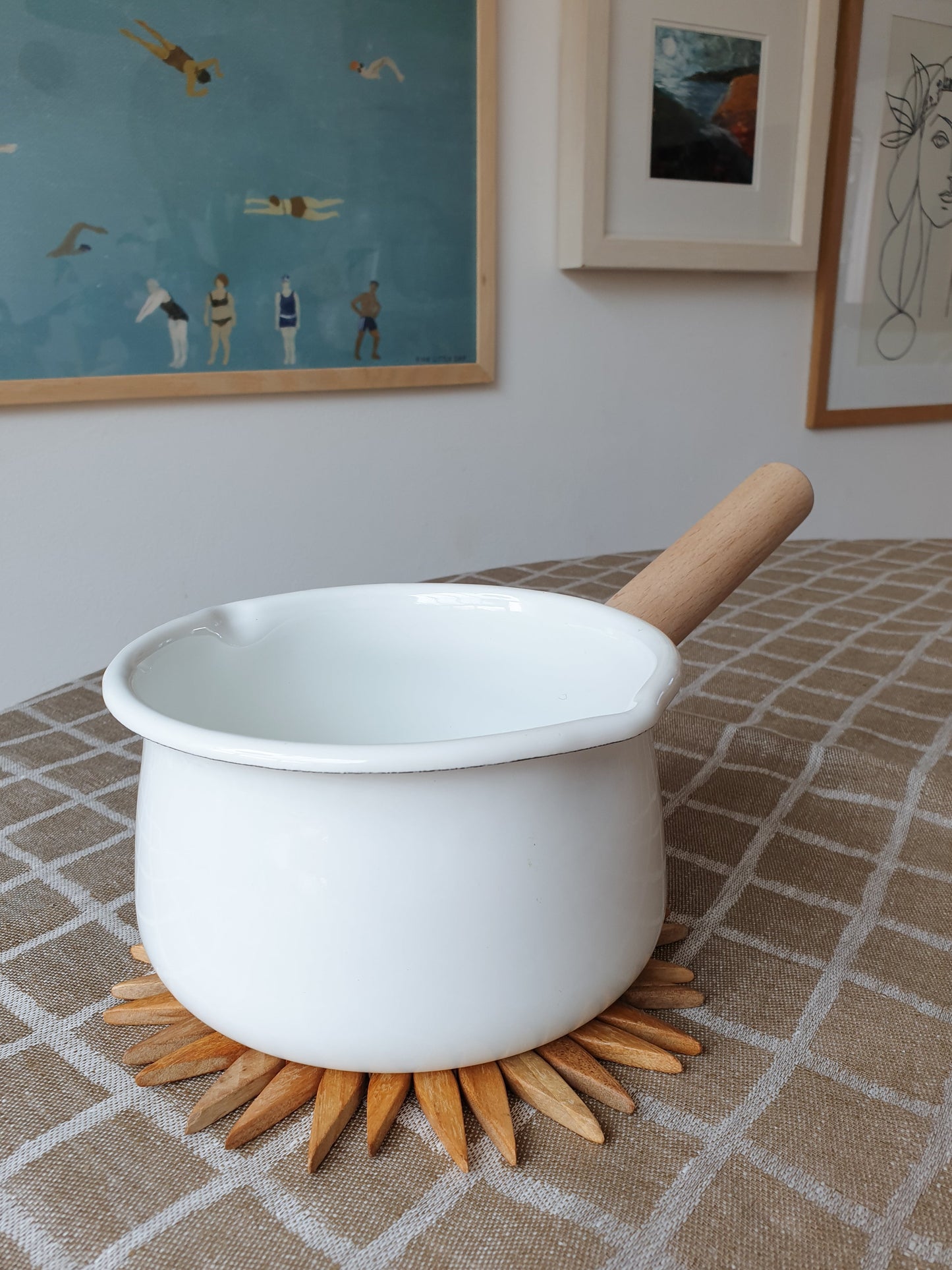 Small two-tone wooden starburst trivet protecting a linen tablecloth under a white enamel saucepan with wooden handle