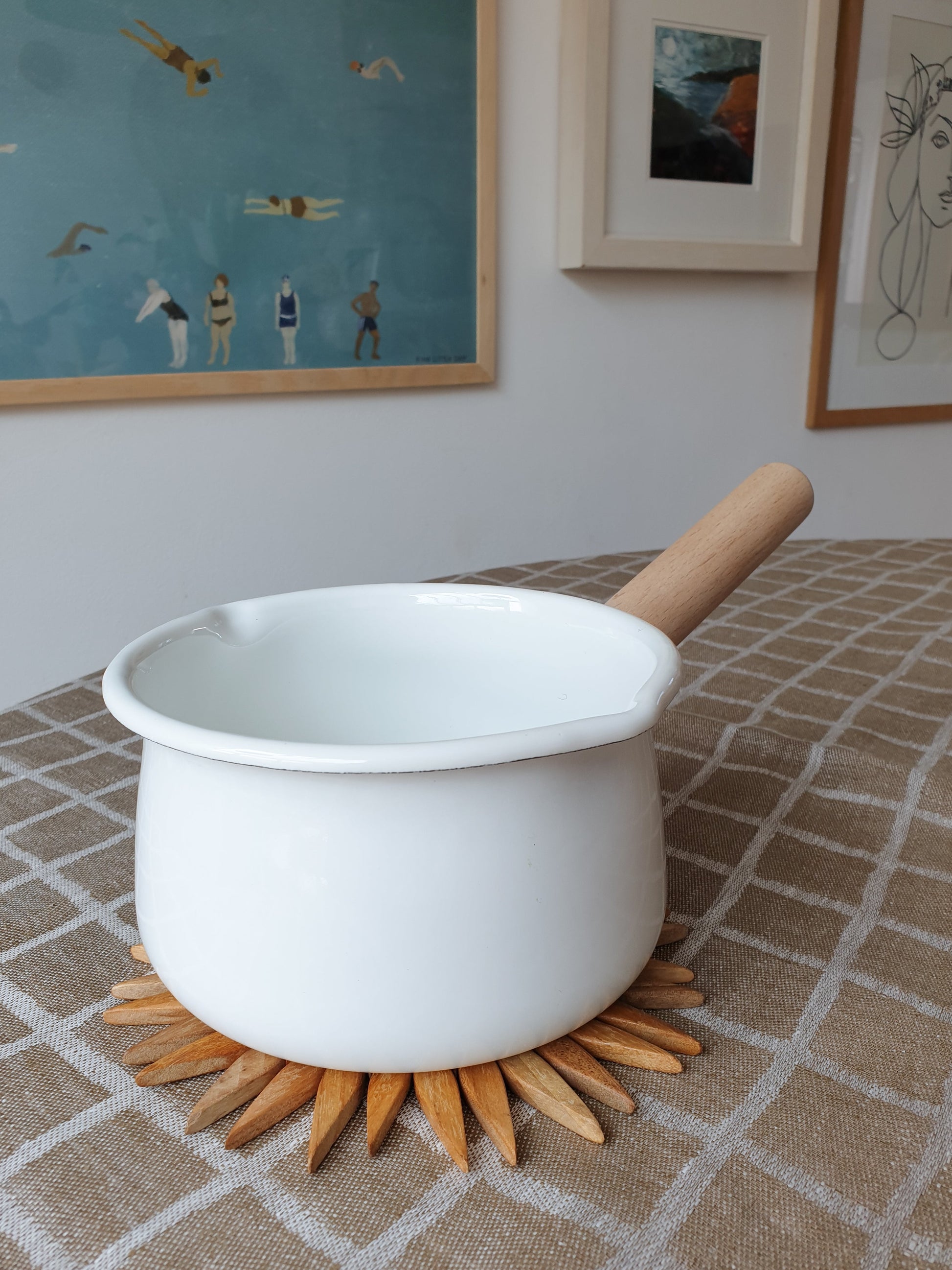 Small two-tone wooden starburst trivet protecting a linen tablecloth under a white enamel saucepan with wooden handle