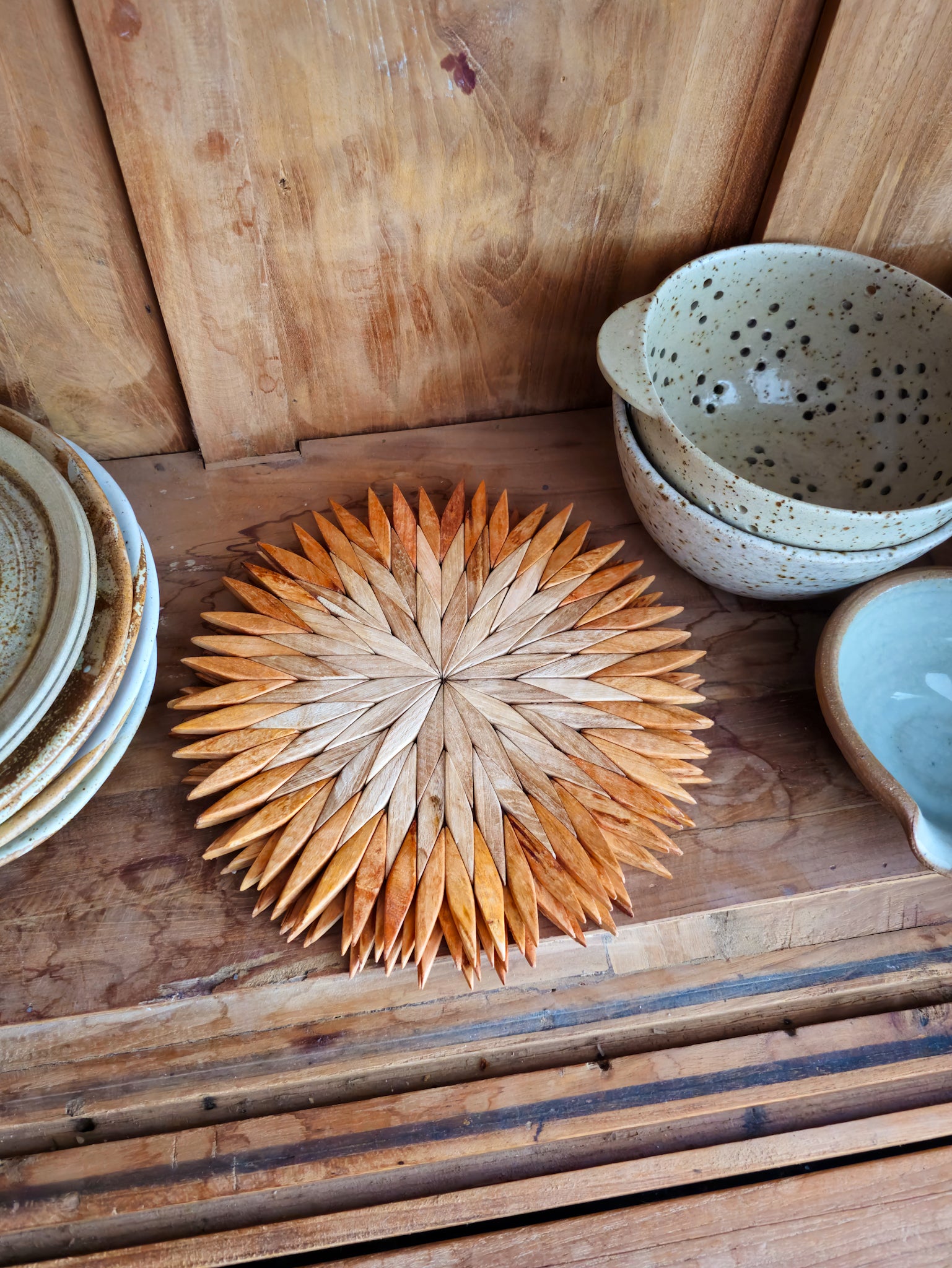 Large two-tone wooden starburst trivet on a wooden kitchen shelf, styled with handmade ceramic bowls and plates
