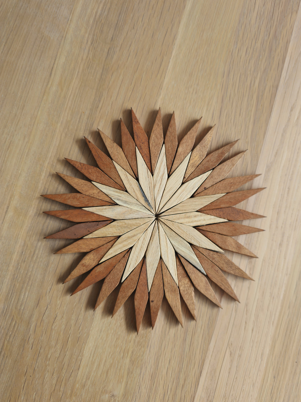 Small two-tone wooden starburst trivet in Carpathian beechwood, with a pale centre and warm brown outer petals, on an oak table