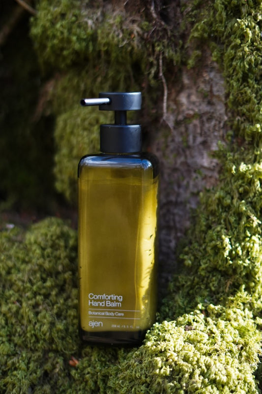 A pump bottle of Ajen Hand Balm placed on a natural surface with a mossy background, indicating the product is a hand balm with a natural scent profile.