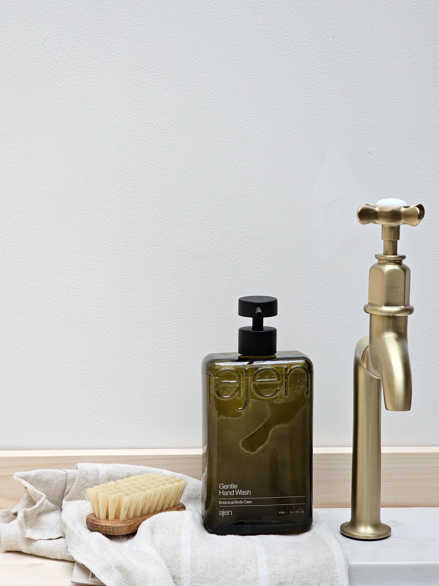 A bottle of Ajen hand wash next to a gold-colored faucet, with a white cloth and a wooden brush in the foreground.