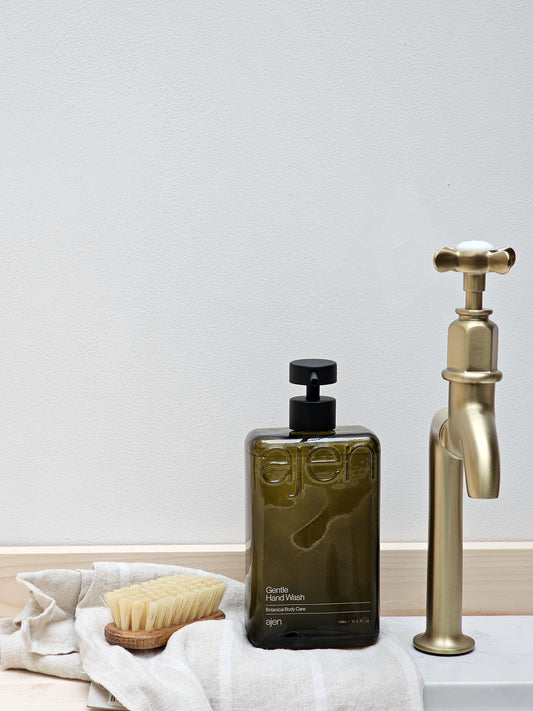 A bottle of Ajen hand wash next to a gold-colored faucet, with a white cloth and a wooden brush in the foreground.